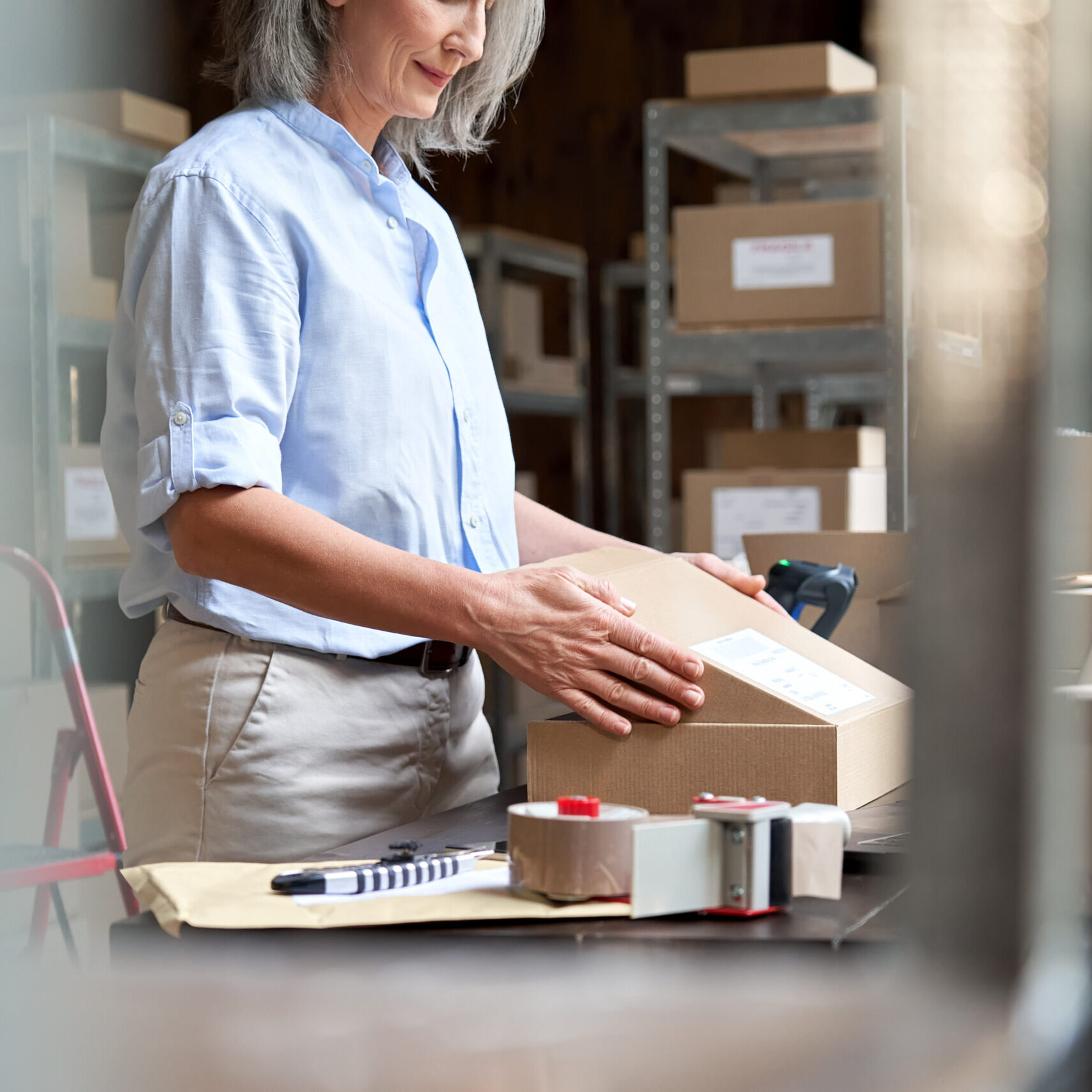 Close up over shoulder view of old female online store small business owner worker packing package post shipping ecommerce retail order in box preparing delivery parcel on table. Dropshipping service.