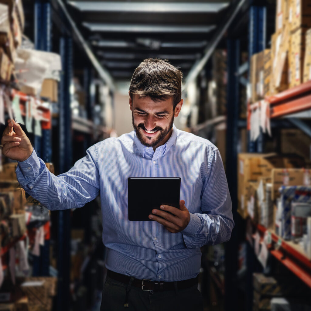 Smiling bearded supervisor standing in warehouse full of boxes and checking on goods ready for deliver.