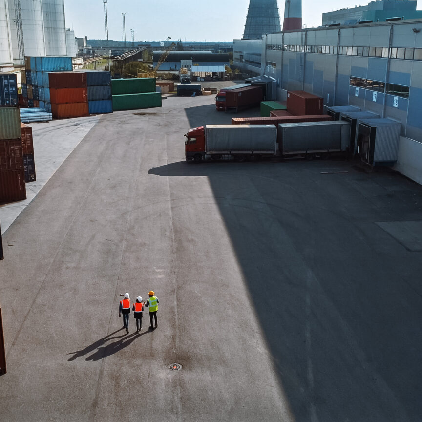 Aerial Shot of a Team of Diverse Industrial Engineers, Safety Supervisors and Foremen in Hard Hats and Safety Vests Walking in Cargo Container Terminal Depot. Colleagues Talk About Logistics.